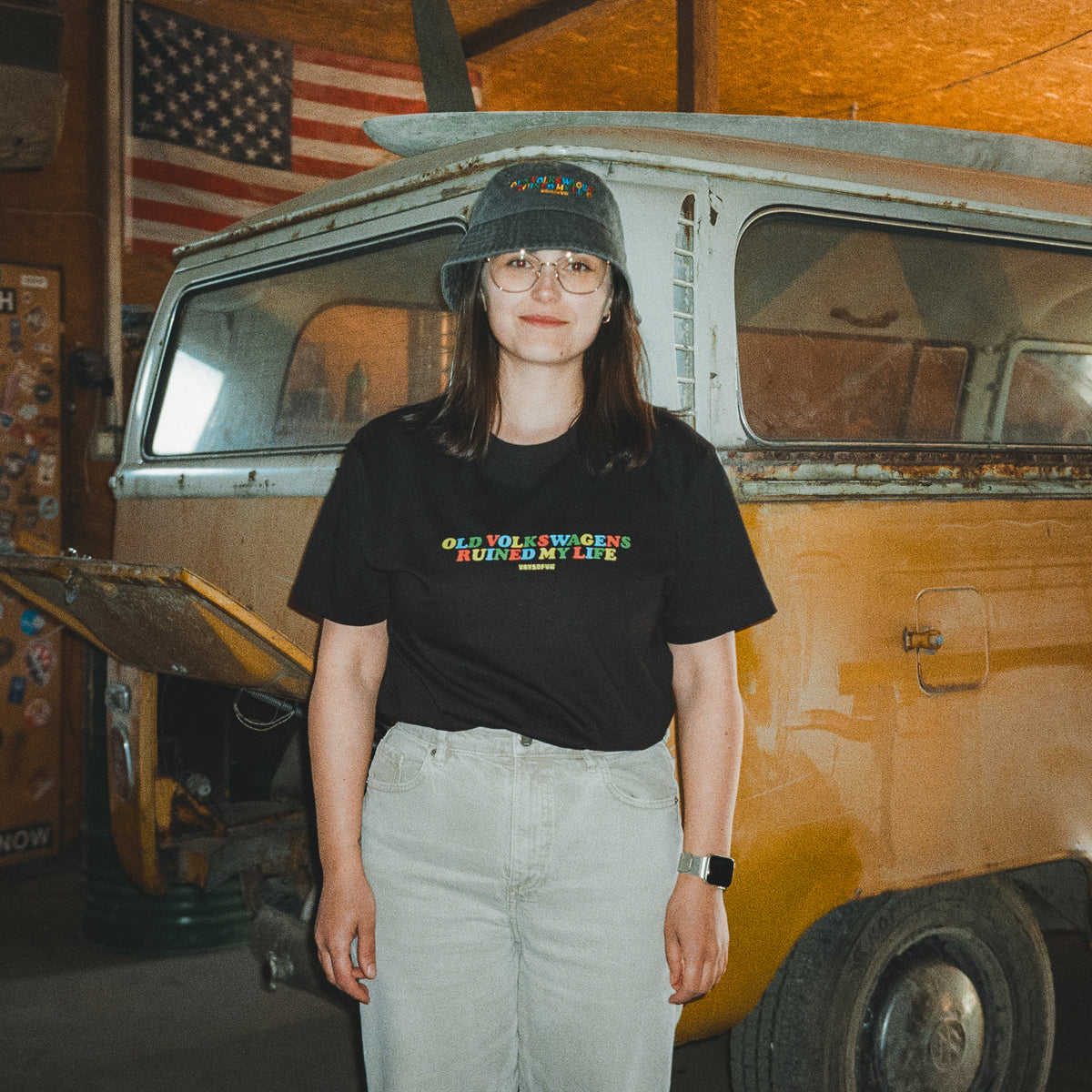Woman wearing black VW T1 Bus tee with Old Volkswagens Ruined My Life print, standing in front of a classic Volkswagen Bus in a garage – vansofvw