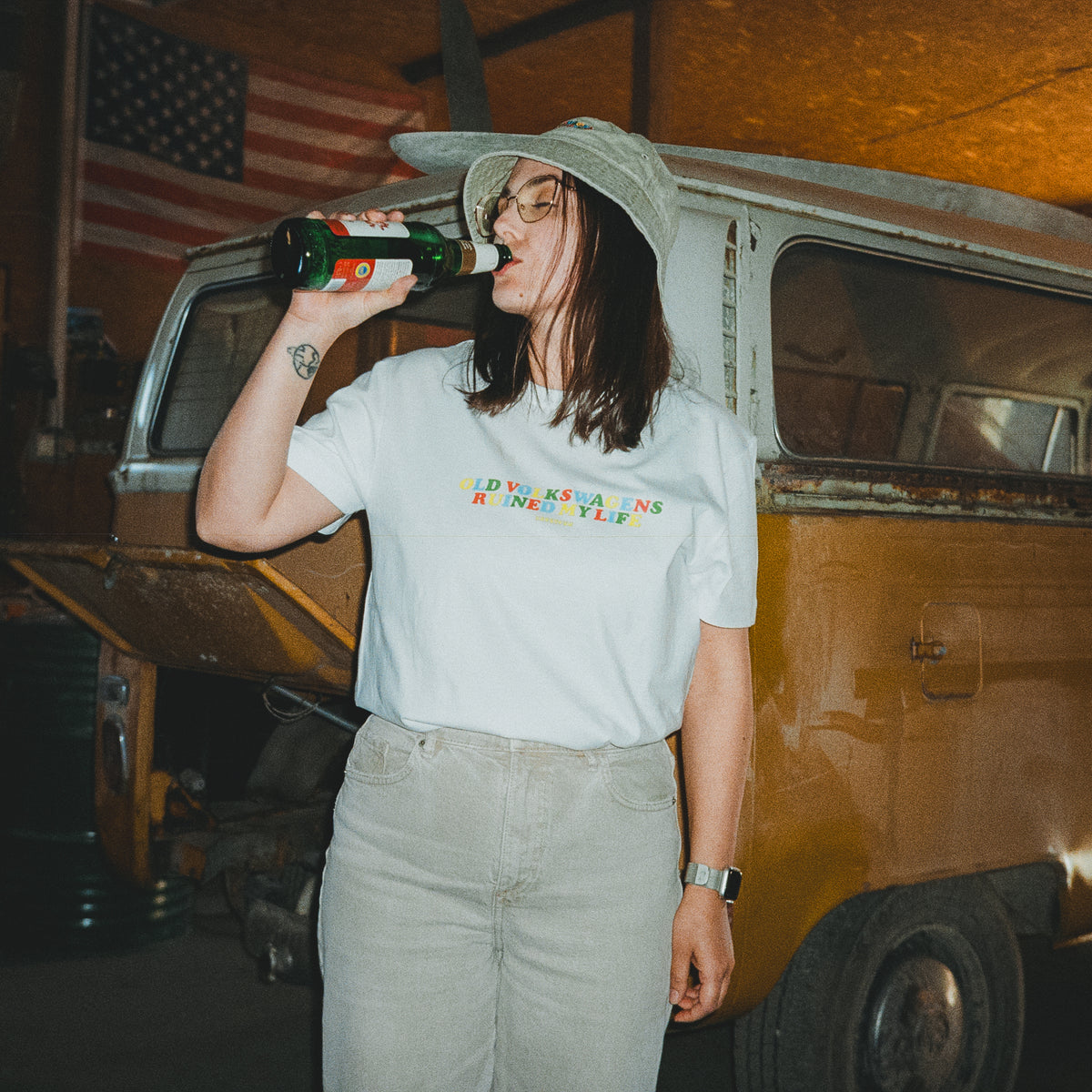 Woman wearing white VW T1 Bus tee with colourful Old Volkswagens Ruined My Life print, in front of a rusty classic Volkswagen Bus – vansofvw