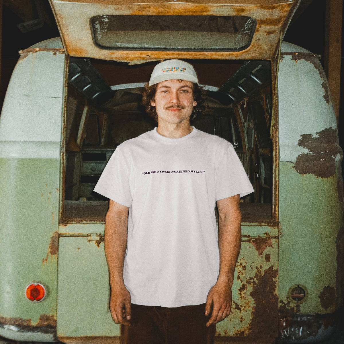 Person wearing a white t-shirt with text standing in front of a vintage, rusted vehicle.